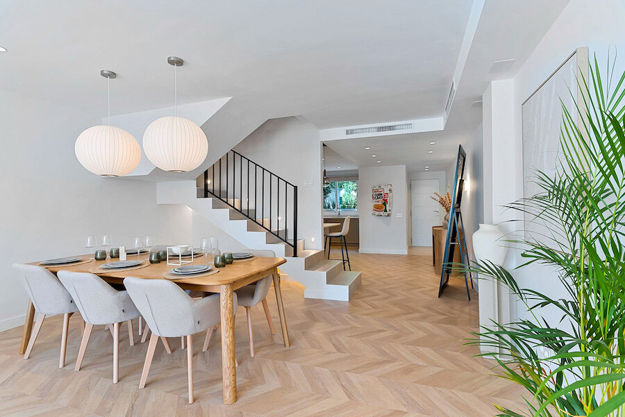 Salle à manger lumineuse à aire ouverte dans une maison mitoyenne du Golden Mile de Marbella, avec table en bois, suspensions modernes, parquet en chevrons et escalier menant à l’étage supérieur.