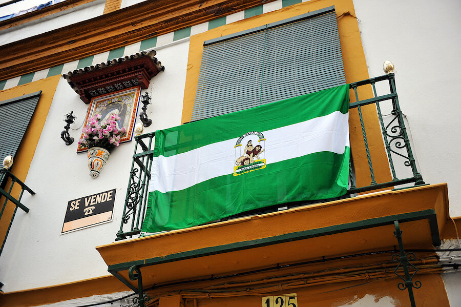 Día de l’Andalousie. Drapeau andalou suspendu au balcon d’un bâtiment traditionnel aux murs blanchis à la chaux avec des encadrements jaunes, doté de balustrades en fer forgé et d’un carreau céramique religieux décoratif sur la façade.