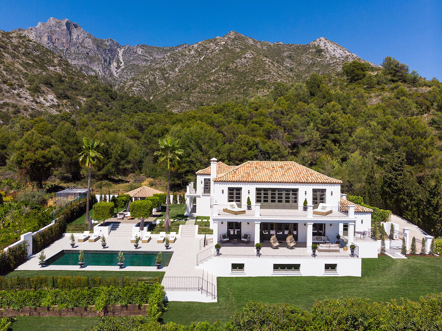 White Mediterranean-style villa with terracotta roof and swimming pool, set in landscaped grounds beneath a dramatic mountain backdrop under a clear blue sky. New on the market