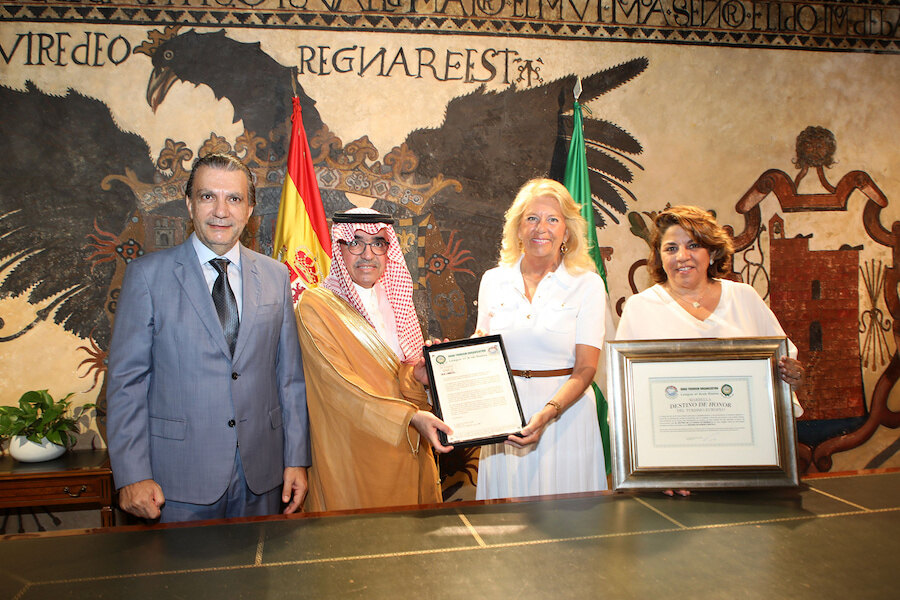 Arab Tourism. Four officials standing in front of Spanish and Saudi flags during a formal ceremony, holding framed certificates and an award plaque, with a historic mural in the background.