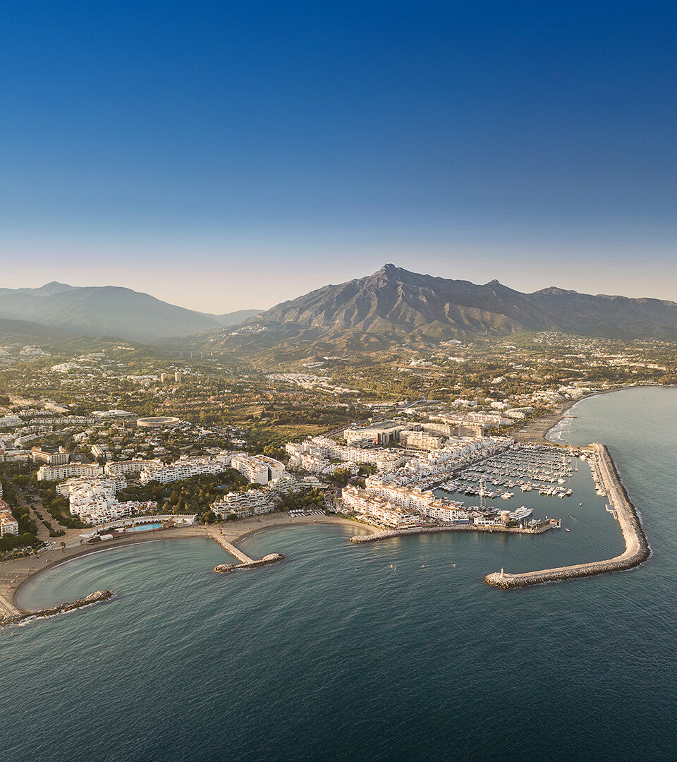Aerial panoramic view of Puerto Banús marina and Marbella’s coastline, with the Mediterranean Sea in the foreground and La Concha mountain under a clear blue sky.