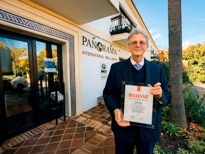 Christopher Clover in a navy suit standing outside the Panorama International Real Estate office in Marbella, holding a “Bellevue Best Property Agents 2026” award plaque, with palm trees and mountains in the background.