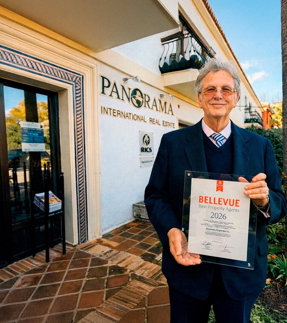 Christopher Clover in a navy suit standing outside the Panorama International Real Estate office in Marbella, holding a “Bellevue Best Property Agents 2026” award plaque, with palm trees and mountains in the background.