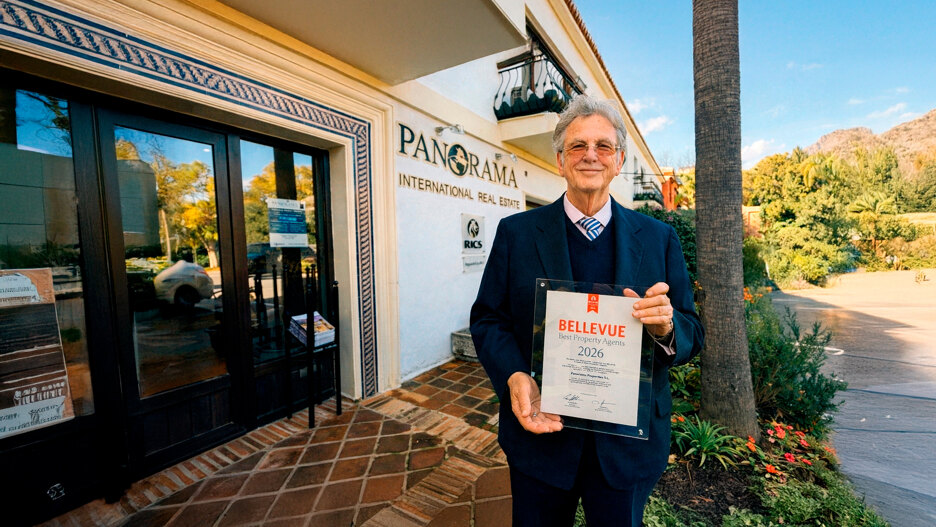 Christopher Clover, con traje azul marino, posando frente a la oficina de Panorama International Real Estate en Marbella, sosteniendo una placa del premio «Mejores Agentes Inmobiliarios Bellevue 2026», con palmeras y montañas al fondo.
