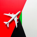 Top view of a white airplane model placed diagonally over a background inspired by the UAE flag, featuring red, green, black, and white geometric sections.
