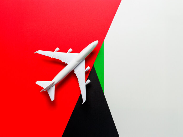 Top view of a white airplane model placed diagonally over a background inspired by the UAE flag, featuring red, green, black, and white geometric sections.