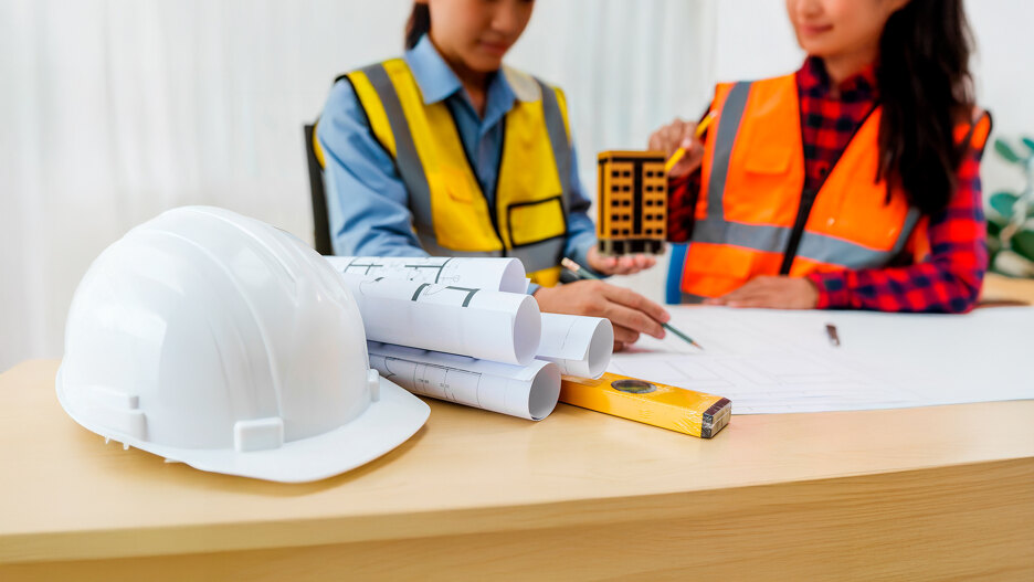 Construction professionals reviewing architectural plans at a desk with blueprints, a safety helmet, measuring tools and a building model.