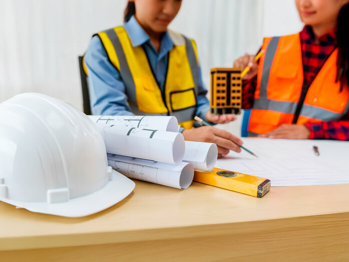 Des professionnels du bâtiment examinant des plans architecturaux sur un bureau, avec des plans, un casque de sécurité, des outils de mesure et une maquette de bâtiment.