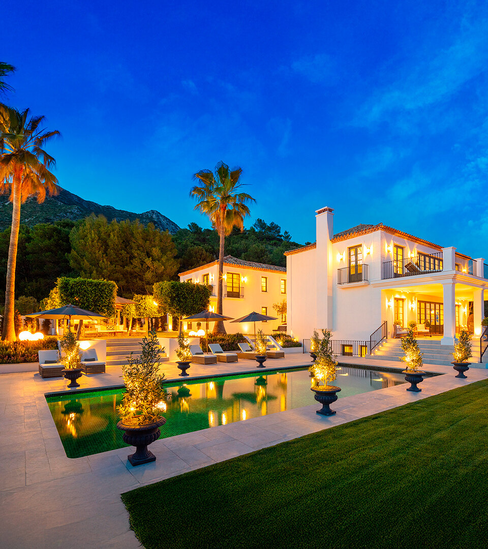 Illuminated luxury villa at dusk with swimming pool, palm trees and landscaped garden, set against mountain backdrop under a deep blue sky.