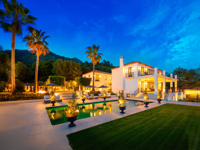 Illuminated luxury villa at dusk with swimming pool, palm trees and landscaped garden, set against mountain backdrop under a deep blue sky.