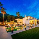 Illuminated luxury villa at dusk with swimming pool, palm trees and landscaped garden, set against mountain backdrop under a deep blue sky.