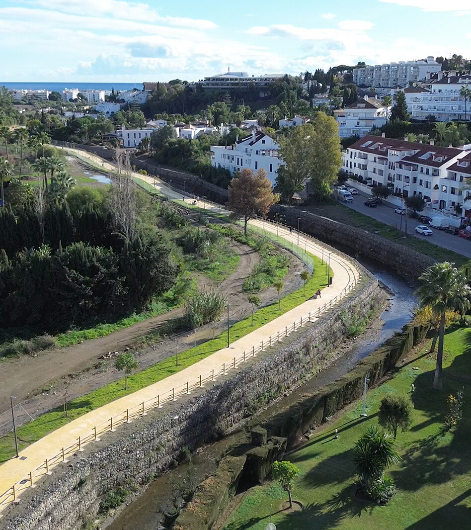 Aerial view of Marbella’s new riverside promenade, with a paved path, railings and nearby homes, and the Mediterranean Sea in the distance.