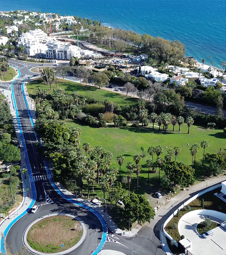 Vista aérea de la zona costera de Marbella, con un parque ajardinado, avenidas bordeadas de palmeras, edificios residenciales modernos y el mar Mediterráneo al fondo.