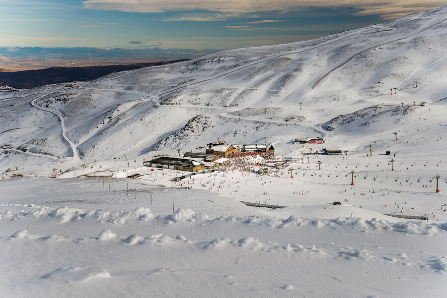 Vue d’ensemble de la base de la station de ski, recouverte de neige, avec des bâtiments, des remontées mécaniques et des skieurs répartis sur les pistes.