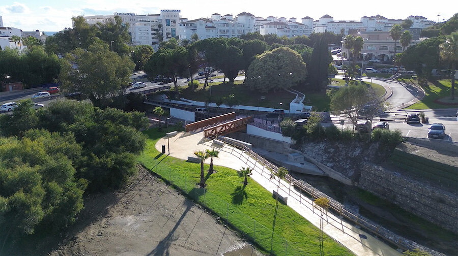 Aerial view of Marbella’s riverside walkway with landscaped lawns, railings and a small footbridge, next to a roundabout and residential buildings.