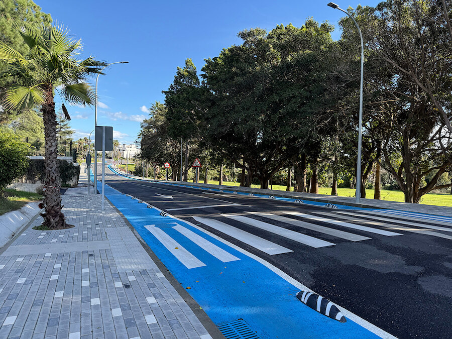 Vista a pie de calle de la carretera de Istán renovada en Marbella, con un carril bici azul segregado, acera peatonal, paso de peatones, palmeras y zonas verdes ajardinadas bajo un cielo despejado.