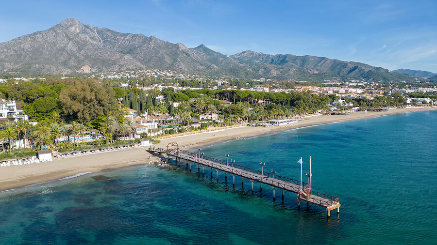 Aerial view of a Marbella beach and pier with turquoise sea in the foreground and La Concha mountain rising behind the coastline.