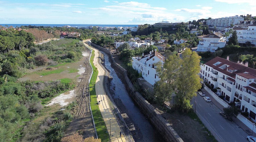 Aerial view of Marbella’s riverside promenade alongside the river channel, with residential buildings on the right and the Mediterranean Sea on the horizon.