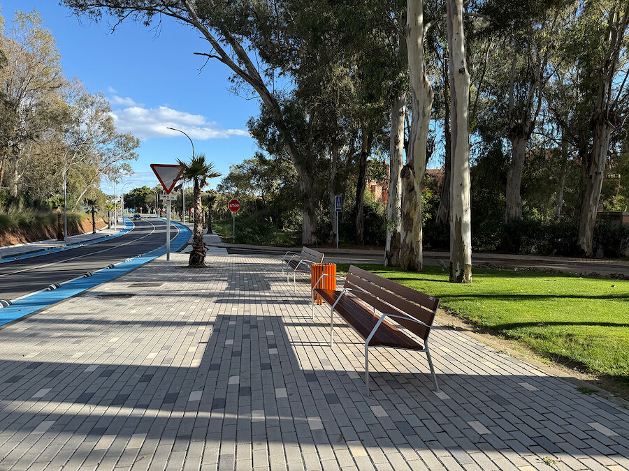 Paseo peatonal junto a una carretera recientemente renovada en Marbella, con bancos, árboles, zonas verdes ajardinadas y un carril bici bajo un cielo despejado.