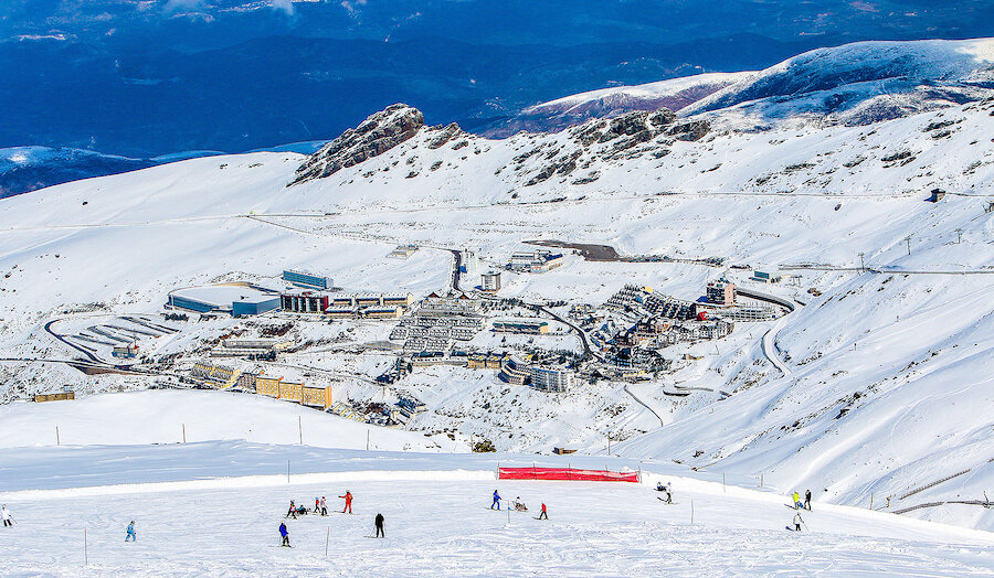 Vue panoramique d’un village de station de ski enneigé en montagne, avec des skieurs sur la piste au premier plan.