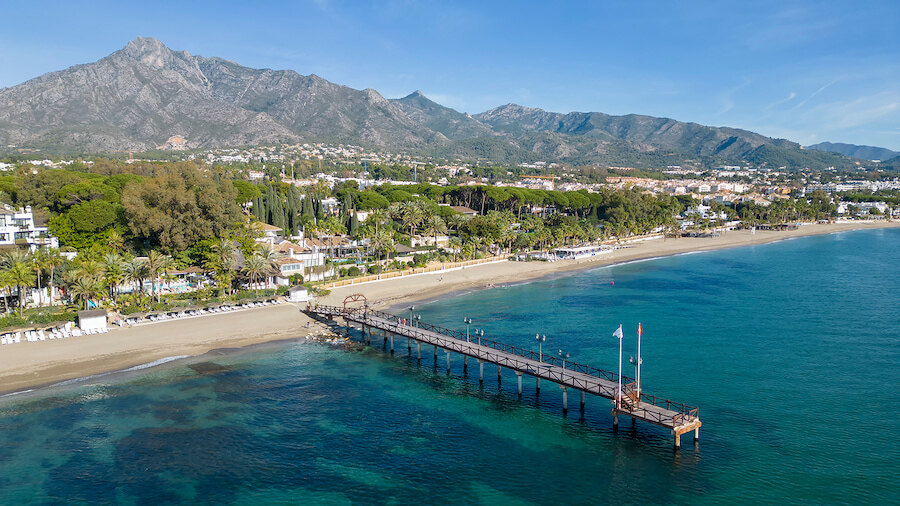 Luftaufnahme der Küste von Marbella mit einem hölzernen Steg, der sich in türkisfarbenes Wasser erstreckt, einem Sandstrand, einer palmengesäumten Promenade und dem Berg La Concha im Hintergrund.