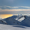 Sommets enneigés surplombant une mer dorée par le soleil et des nuages ondulants, sous un ciel bleu limpide.