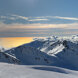 Picos montañosos cubiertos de nieve con vistas a un mar dorado por la luz del sol y nubes ondulantes bajo un cielo azul despejado.