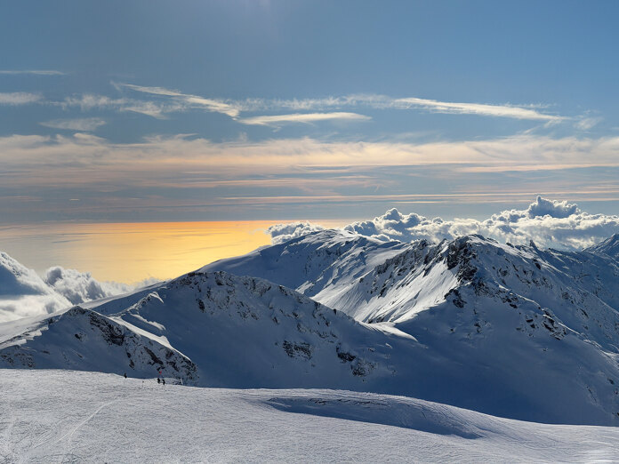 Snow-covered mountain peaks overlooking a golden sunlit sea and rolling clouds beneath a clear blue sky.