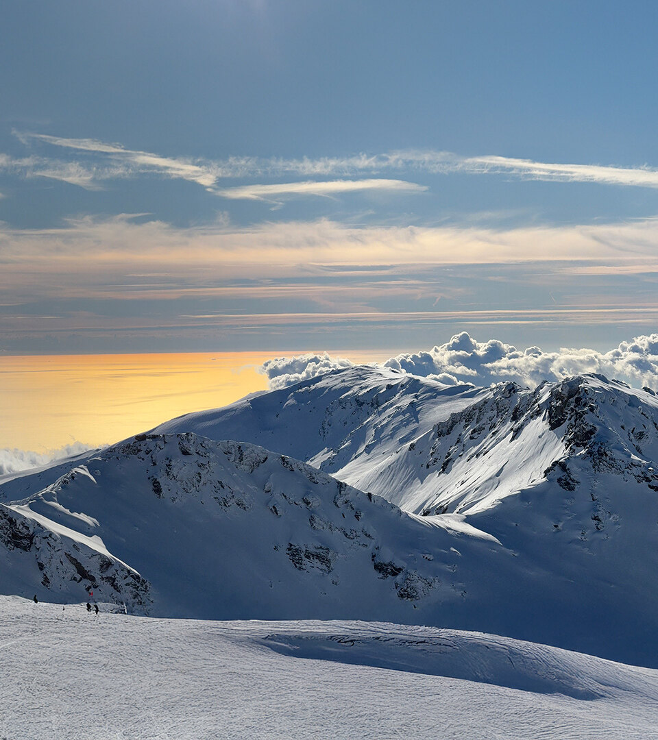 Sommets enneigés surplombant une mer dorée par le soleil et des nuages ondulants, sous un ciel bleu limpide.