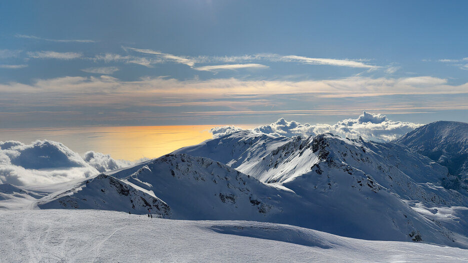 Snow-covered mountain peaks overlooking a golden sunlit sea and rolling clouds beneath a clear blue sky.