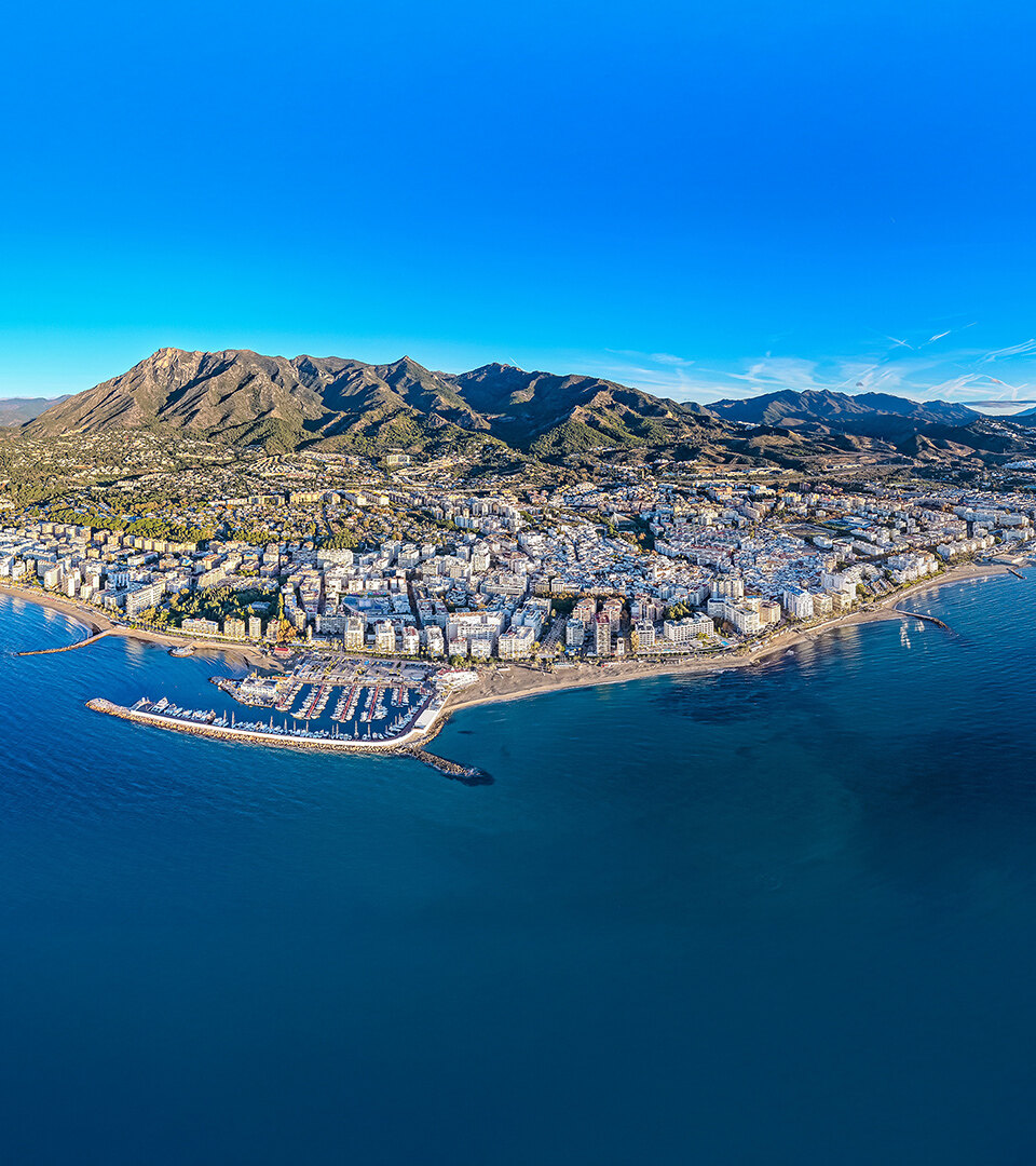 Aerial panoramic view of Marbella’s coastline with the city, marina and mountains behind, bordered by the Mediterranean under a clear blue sky.