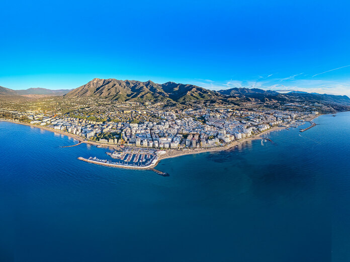 Aerial panoramic view of Marbella’s coastline with the city, marina and mountains behind, bordered by the Mediterranean under a clear blue sky.
