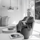 Retrato en blanco y negro de un hombre con traje sentado en una sala de estar de oficina moderna, con imágenes de propiedades enmarcadas y premios del sector expuestos en la pared detrás de él.
