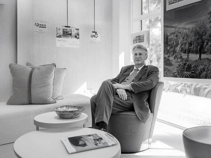 Black-and-white portrait of a suited man seated in a modern office lounge, with framed property images and industry awards displayed on the wall behind him.