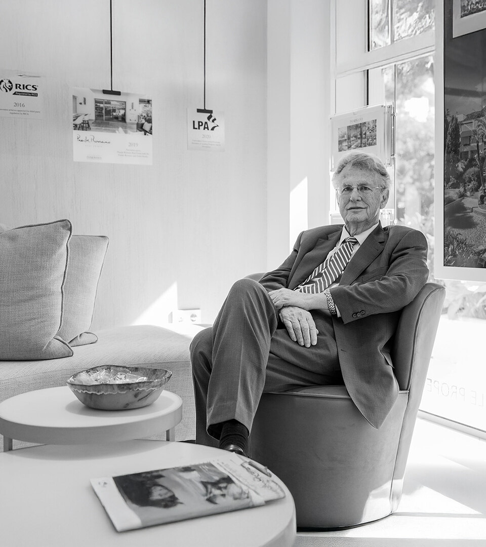Retrato en blanco y negro de un hombre con traje sentado en una sala de estar de oficina moderna, con imágenes de propiedades enmarcadas y premios del sector expuestos en la pared detrás de él.
