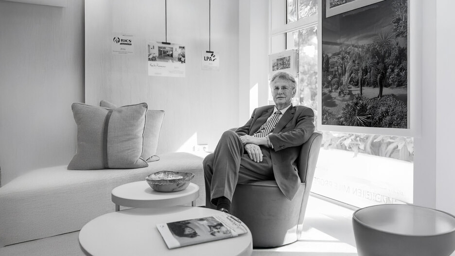 Zwart-witportret van een man in pak, zittend in een moderne kantoorlounge, met ingelijste vastgoedafbeeldingen en brancheprijzen aan de muur achter hem.