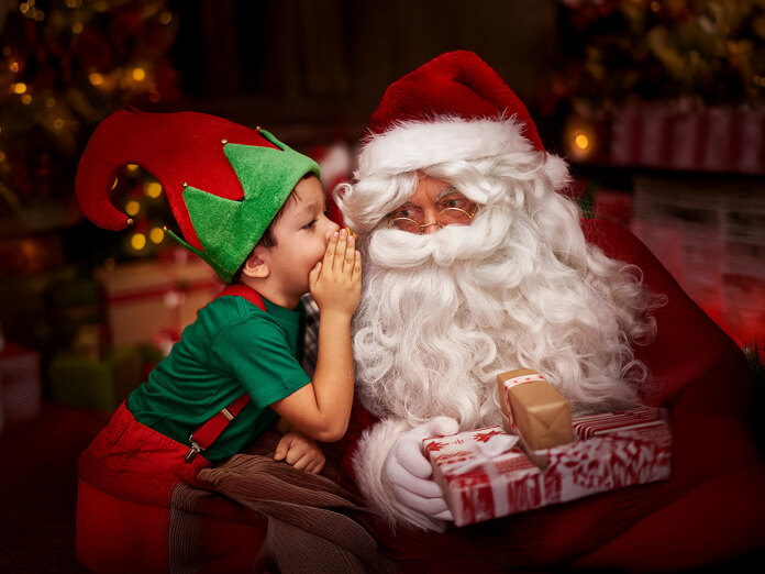 Smiling child dressed as an elf whispering a secret to Santa Claus while he holds wrapped Christmas presents.