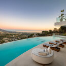 Infinity pool terrace with striped sun loungers and curved lounge chair overlooking hillside homes, mountains and Mediterranean Sea at sunset.