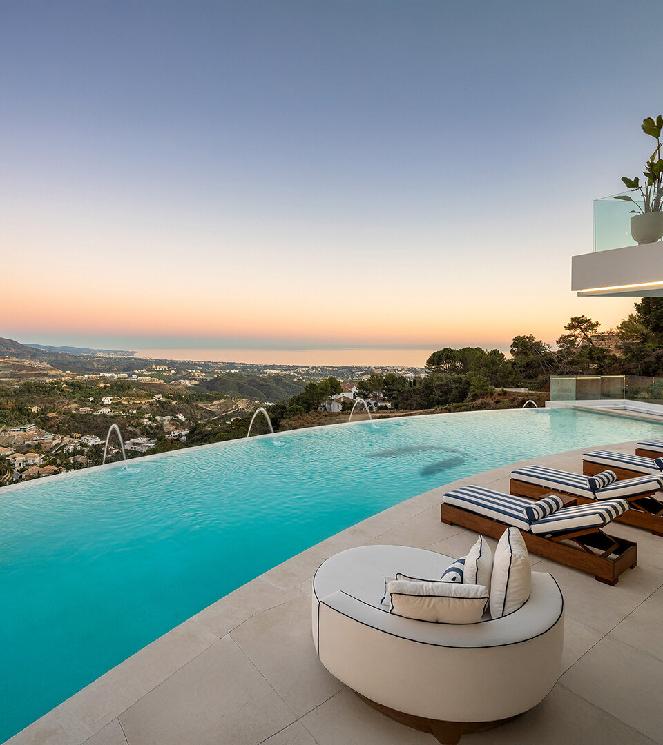 Infinity pool terrace with striped sun loungers and curved lounge chair overlooking hillside homes, mountains and Mediterranean Sea at sunset.