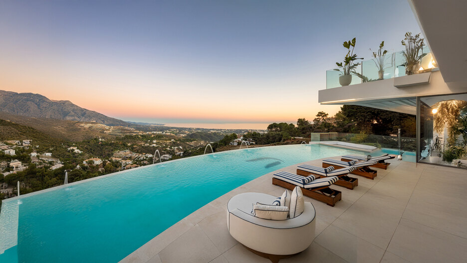 Terraza con piscina infinita, tumbonas a rayas y sillón curvo con vistas a casas en la ladera, montañas y al mar Mediterráneo al atardecer.