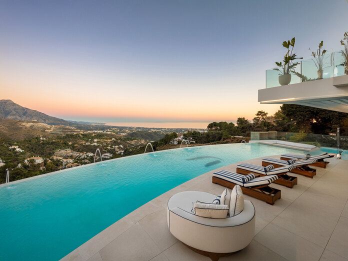 Infinitypool-terras met gestreepte ligstoelen en een gebogen loungestoel, uitkijkend over heuvelvilla’s, bergen en de Middellandse Zee bij zonsondergang.