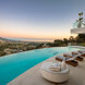 Infinity pool terrace with striped sun loungers and curved lounge chair overlooking hillside homes, mountains and Mediterranean Sea at sunset.