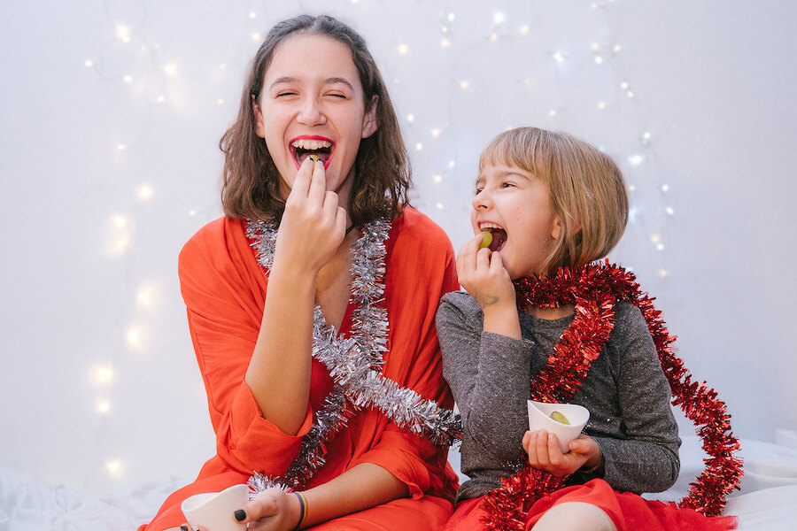 Two laughing children wrapped in Christmas tinsel, eating grapes from small bowls in front of festive fairy lights.