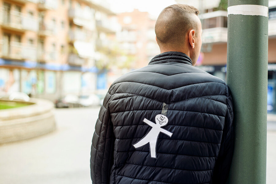 Man wearing a black jacket in a city street with a paper stick figure taped to his back as a playful prank.
