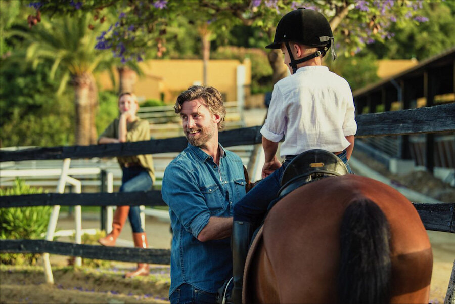 Smiling man assisting a young child on horseback at an equestrian arena, with a woman watching in the background among palm trees.