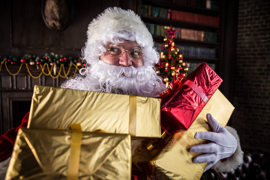 Santa Claus holding a stack of gold and red wrapped Christmas presents in a cosy, festively decorated room.
