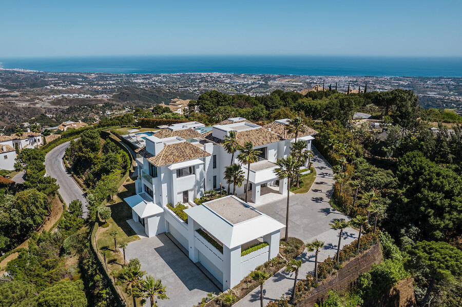 Aerial view of a contemporary white hillside villa with lush gardens, long driveway and panoramic sea and coastline views.