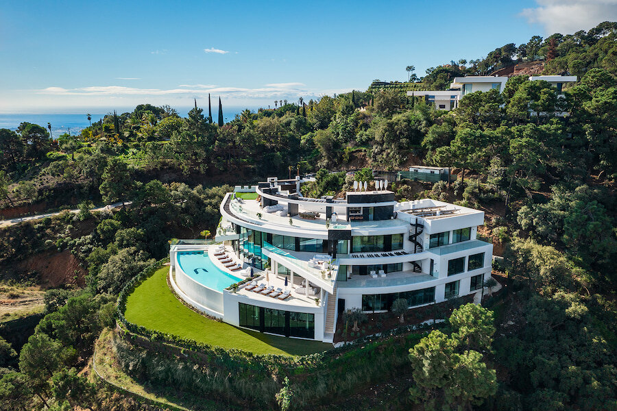Aerial view of a curved, ultra-modern white hillside villa with infinity pool, lawn and wide terraces overlooking a forested valley and the Mediterranean Sea. la zagaleta