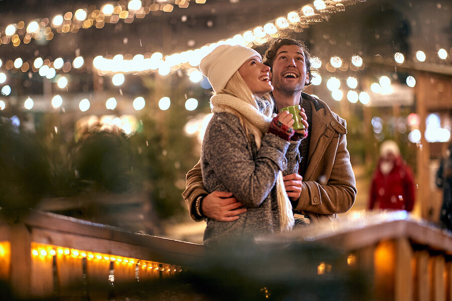 Smiling couple in winter clothes enjoying a hot drink together at an outdoor Christmas market with festive string lights.