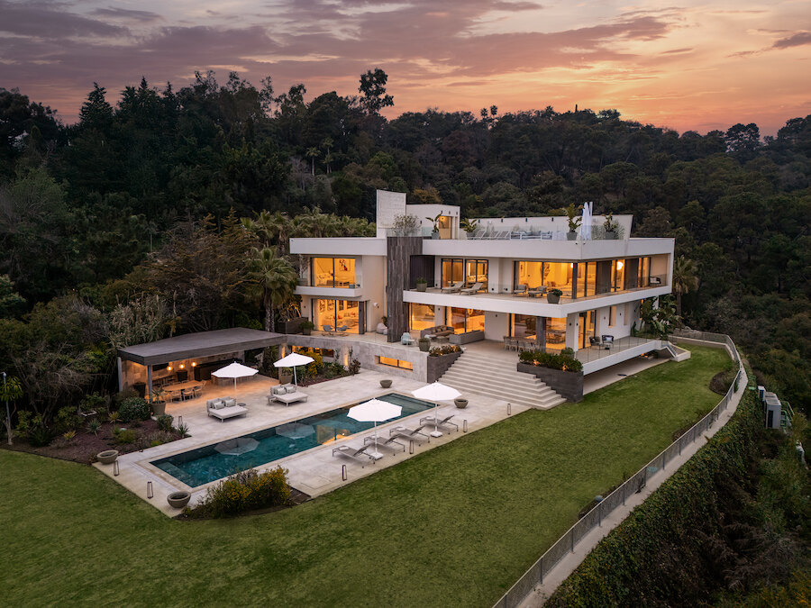 Modern hillside villa at sunset with expansive terraces, illuminated interiors, lawn, pool and poolside lounge areas overlooking woodland.